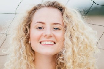 Close up shot of curly lovely young female has broad shining smile, looks positively at camera, has joyful expression, stands outdoor, has fresh skin, being glad to be photographed. Headshot