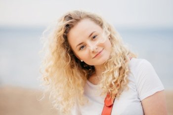 Outdoor shot of curly beautiful young female with fresh skin, looks positively at camera, wears casual white t shirt, poses against blurred seaside background. People and recreation concept.