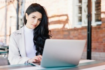 Portrait of beautiful brunette female freelancer typing a message via email using laptop while sitting on terrace looking at camera with confident look. Young lady working with computer outdoors