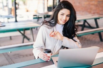 Glad young self-employed brunette woman enjoying free wireless internet connection sitting in front of generic laptop at outdoor cafe holding takeaway coffee. Relaxation, beauty and youth concept