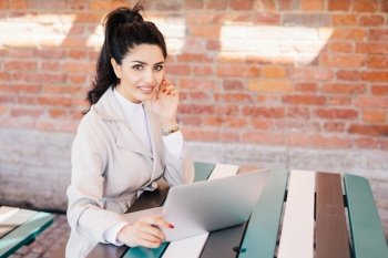 Succsessful businesswoman with dark hair, nice eyes, healthy skin, well-shaped lips having red long nails wearing formal clothes while sitting at cafe using laptop for online communication and work