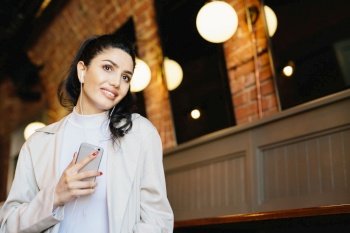 Caucasian beautiful woman with dark hair, appealing big eyes with long eyelashes and full lips having long fingers holding mobile phone in hand listening to music with earphones standing indoors