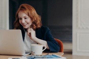 Happy smiling female entrepreneur has online conference via modern laptop computer, connected to wireless internet at home, uses earphones to hear interlocutor better, sits in coworking space