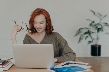 Busy redhead adult woman does freelance work on laptop computer, focused in monitor, prepares project, surrounded with paper documents, wears warm sweater, analyzes information, uses internet