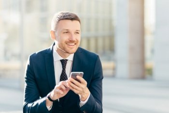 Delighted thoughtful elegant male marketing trader in formal black suit, uses modern cell phone for searching website in internet, types text message, looks pensively into distance. Bussiness concept