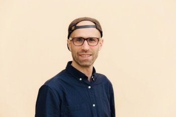 Portrait of satisfied Caucasian male with stubble, has gentle smile, wears stylish cap and shirt, looks through spectacles, poses against beige background. Hipster guy being in good mood. Horizontal