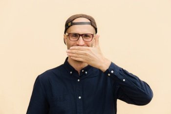 Positive male model covers mouth as giggles at funny joke, wears spectacles, cap and elegant shirt, expresses good emotions, poses against beige background. People, emotions, lifestyle concept