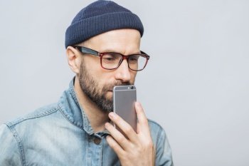 Thoughtful middle aged male with stubble holds smart phone near mouth, being deep in thoughts, thinks about future plans, isolated over white background. People, emotions, technology concept