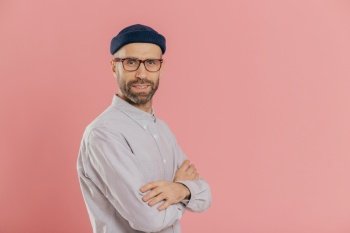Studio shot of self assured bearded man with blue eyes, wears stylish hat and white shirt, keeps arms folded, models over pink background with free space on left side for your advertising content