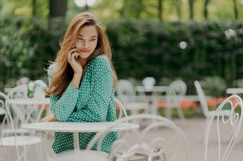 Outside shot of charming young woman with long hair, wears polka dot green shirt, sits at table in outdoor cafe, has pleasant talk via modern smartphone, has dreamy expreession. People and lifestyle