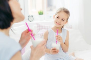 Cropped shot of beautiful small female child drinks tasty milk shake together with her mother, enjoys good morning and calm domestic atmosphere, has happy look. People, breakfast and bedding