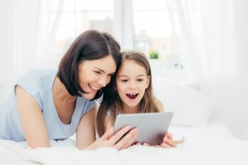 Positive cheerful small female kid and her young mother watch interesting cartoon on tablet, connected to wireless internet, pose against bedroom interior, have happy expressions. Family concept