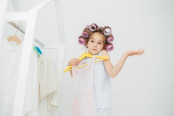 Hesitant girl with clueless expression, doesn`t know what to wear, holds hangers with dress, has curlers on head, isolated over white background. Cute child with pleasant appearance prepares for party