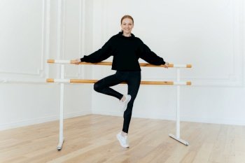 Horizontal shot of flexible young female leans on ballet barre, wears black costume and white sportshoes, smiles positively, poses in dancing hall. Ballet instructor demonstrates different exercises