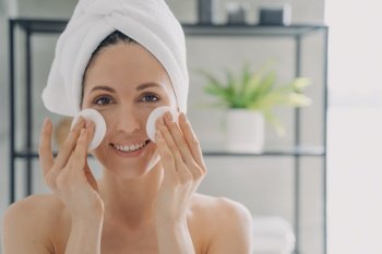 Girl is cleansing skin with face toner and cotton disks. Attractive caucasian woman wrapped in towel after bathing. Young woman takes shower at home. Delicate skin care and rejuvenation.. Girl is cleansing skin with face toner and cotton disks. Attractive young woman after bathing.