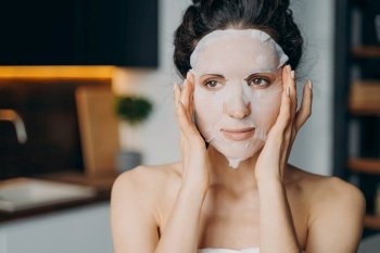 Caucasian girl applies mask sheet after bathing. Vitamin, nourishing and nutrition face mask. Young woman takes shower at home or hotel and doing spa procedures. Skin care and rejuvenation.. Caucasian girl applies mask sheet after bathing. Vitamin, nourishing and nutrition face mask.