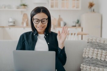 Handsome spanish girl in glasses has video call on laptop and waving hand. Woman says hello to business partner. Remote work on quarantine. Businesswoman is communicating with colleagues for project.. Handsome girl in glasses has video call and waving hand. Communicating with colleagues for project.
