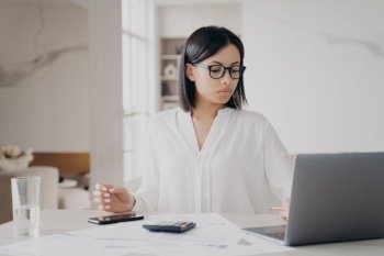 Concentrated european businesswoman in glasses and white blouse is working remote from home. Manager, accountant or business assistant is doing paperwork using laptop. Success and career concept.. Concentrated european businesswoman is working remote from home. Success and career concept.