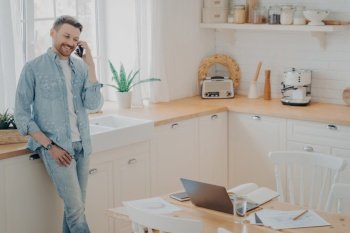 Happy young caucasian man holding cellphone while having conversation with client, standing in modern kitchen and enjoying good business talk while working distantly from home. Happy young caucasian man having conversation on cellphone in kitchen