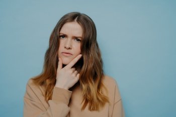 Puzzled young girl with thick wavy ombre hair demonstrating concern and interest while holding hand at chin wearing casual light brown sweater and standing against blue studio background. Confused young woman looking at camera standing over blue background