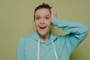 Surprised young woman without makeup demonstrating shock and excitement holding hand at head looking at camera wearing casual blue hooded sweater while posing in studio. Human emotions concept. Excited young woman expressing shock holding hand at head