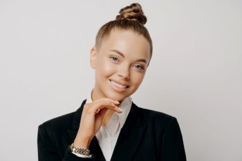 Close up studio portrait of beautiful female business worker with hair in bun wearing formal suit with white shirt, smiling with confidence, putting hand on chin, isolated on grey background. Attractive female business worker smiling at camera