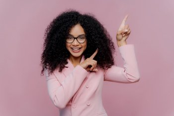 Half length shot of cheerful Afro American woman with dark bushy hairstyle, wears violet suit, points above with both index fingers, demonstrates way to something, invites you going upstairs