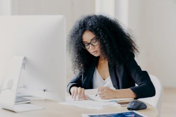 Photo of serious Afro woman writes in papers, sits at table with modern computer, creats article in newspaper, wears glasses and eyewear, poses indoor, works remotely. People and job concept