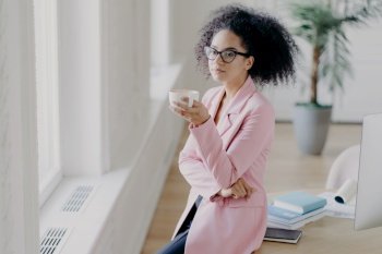 Indoor shot of thoughtful curly haired young woman holds cup of coffee, has pensive look at window, poses in coworking space, leans at table, being in office, dressed elegantly, wears spectacles