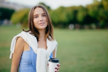 People, recreation and lifestyle concept. Beautiful young European woman has serious look at camera, dressed casually, spends free time at nature, drinks hot beverage, enjoys rest, peaceful atmosphere
