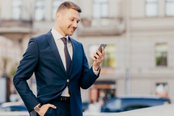 Horizontal shot of happy businessman keeps hand in pocket, wears formal suit and wristwatch, recieves notification on smart phone, finds out about high financial balance. Cheerful trader outdoor