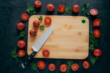 Tomatoes frame around wooden cutting board. Ripe vegetables and slice, green parsley and dill near kitchen board and knife. Food art