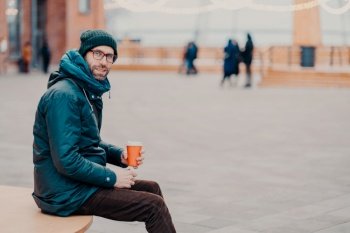 Sideways shot of handsome male adult dressed in street clothes, enjoys aromatic beverage, poses outdoor against blurred background with copy space for your promotion. People and rest concept