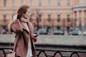 Outdoor shot of relaxed young female looks aside with thoughtful expression, holds modern cell phone, waits for call, dressed in white sweater and coat, carries bag, stand on bridge in big city