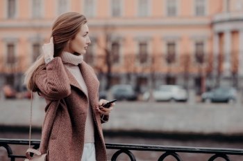 Photo of attractive young female model with long hair, dressed in ncoat, has outdoor stroll during autumn time in big city, holds modern cell phone, text messages, admires beautiful cityscape
