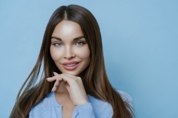 Isolated shot of attractive brunette woman with calm expression, healthy skin and makeup, keeps hand under chin, cares about complexion, looks beautiful, isolated on blue background, models in studio