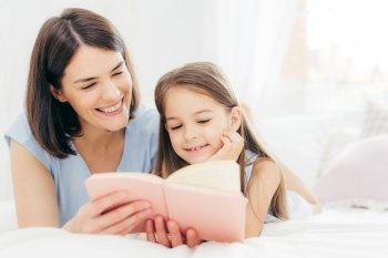 Pretty female mother and her small daughter read interesting book in bedroom, lie on comfortabled bed during morning. Lovely mum reads fairty tale for her little female kid, who is very curious