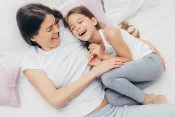 Photo of adorable small child and her mother have fun together in bed, tickle each other, smile joyfully, play after good sleep, have good relationships. People, family, motherhood and childhood
