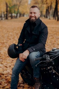 Vertical image of smiling motorcyclist smiles happily, holds helmet, poses on motorbike, wears black jacket and jeans, has drive through park, enjoys autumn time rides black vehicle. Transport concept