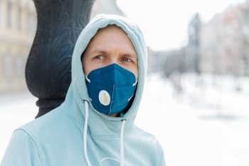 Serious pensive man wears hoodie and protective respiratory mask while walk on street, protects against infection with influenza virus or coronavirus, avoids public crowded places, cares about health