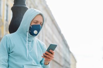 Man on street wears respiratory mask, walks through city, uses modern mobile phone concentrated into display uses wireless internet prevents himself from coronavirus infection. Rapidly spreading virus