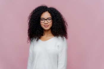 Half length shot of attractive African American woman looks through transparent glasses, white sweater, has serious calm expression, poses against violet studio wall. Facial expressions concept