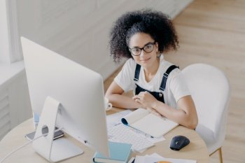 Top view of delighted curly female worker poses at desktop, dressed in casual wear, works on computer, makes financial report. Afro American student prepares for final exam in coworking space