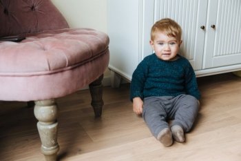 Adorable toddler with charming blue eyes and blonde hair, sits on floor against home interior, plays alone. Sweet baby boy looks directly innto camera, has pleasant appearance. Childhood concept
