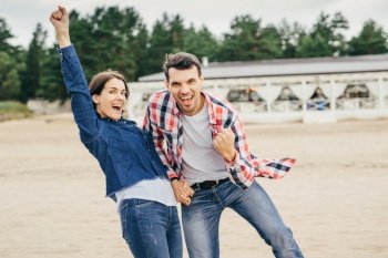 Joyful couple have fun together, clench fists, rejoice their success, have good mood, have happy expressions, holds hands together, pose outdoor. People, happiness and positiveness concept. Joyful couple have fun