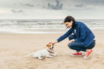 Brunette stylish woman teases her small dog on sea shore, have walk together, have real friendship, understand each other. Beautiful young female spends free time with pet outdoors. Dog and woman on seashore