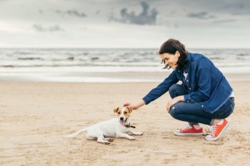 Fashionable woman wears denim stylish clothes and sport shoes spends free time with dog, walk on seashore, enjoy beautiful nature and landscapes. Outdoor shot of female and small pet. Friendship between woman and dog