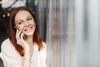 Headshot of smiling female employee with cheerful facial expression, uses smart phone gadget for discussion information with friend, makes mobile conversation, wears formal clothes, stands indoor