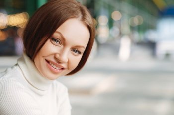 Close up shot of satisfied European woman with cheerful expression, dressed in casual clothes, looks mysteriously at camera, has outdoor walk, poses at street over blurred background with free space