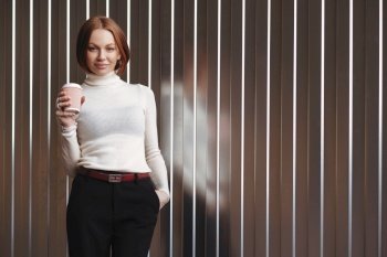 Half length shot of pleasant looking businesswoman dressed in formal clothes, keeps hand in pocket, drinks takeaway coffee, stands against brown wall with free space on left for your advertisement